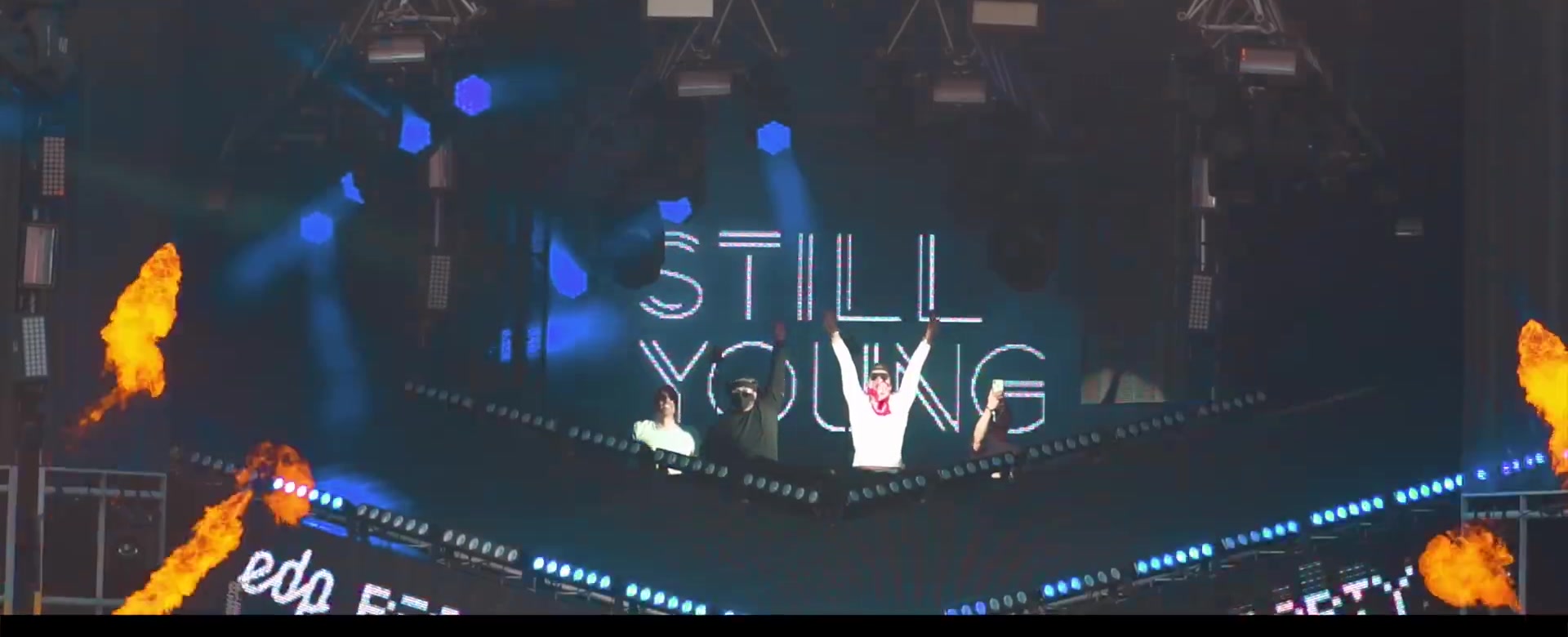 Still Young on a main stage, arms raised beneath the logotype LED with pyro